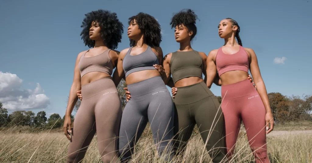 Four women stand together in a grassy field, wearing coordinated seamless athletic sets in earthy tones. They pose confidently under a clear blue sky, gazing into the distance.